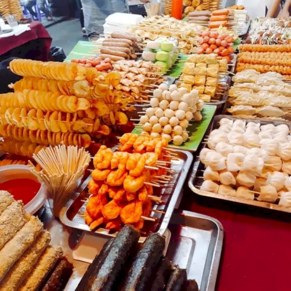 Sizzling grilled skewers, steaming bowls of pho, and fresh tropical fruits under colorful market lights